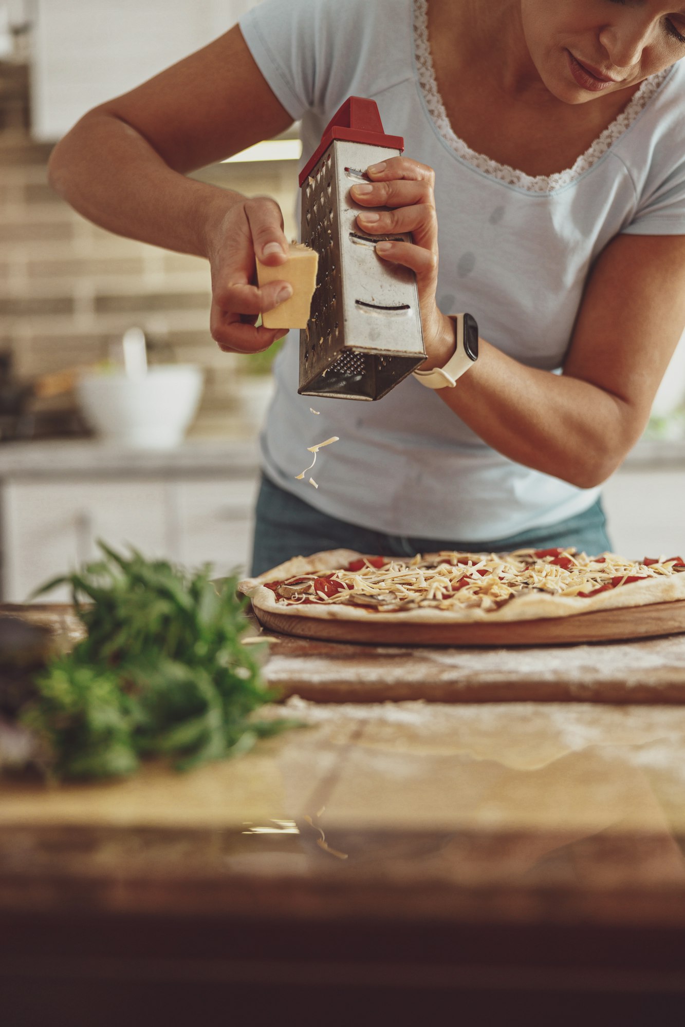 A young housewife cooks pizza adding grated cheese standing in the kitchen