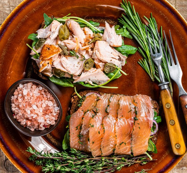 Bruschetta with hot and cold smoked salmon, arugula, capers on a rustic plate with herbs. wooden background. Top view.