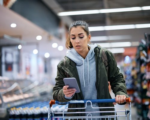 Young woman using shopping app on mobile phone while buying in supermarket.